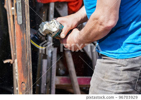 Man polishing wooden door with old angle grinder during sunny day, closeup detail to hands without gloves, fine wood dust flying in air Man polishing wooden door with old angle grinder during sunny day, closeup detail to hands without gloves, fine wood dust flying in air 96703929