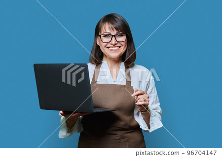 Portrait of middle aged woman in aapron with laptop on blue background. Portrait of middle aged woman in aapron with laptop on blue background. 96704147