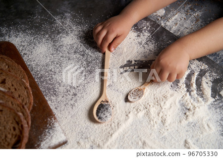 A small child helps to bake.Children's hands hold wooden spoons with seeds.High angle, dark background. Scattered flour and sliced bread in a horizontal photo. Copy space. 96705330