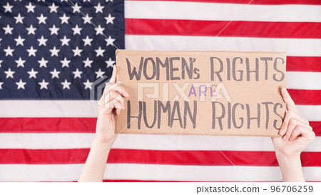 Young woman protester holds cardboard with Womens Rights Are Human Rights sign against USA flag on background. Feminist power. Equal opportunity Womens rights freedom. Young woman protester holds cardboard with Womens Rights Are Human Rights sign against USA flag on background. Feminist power. Equal opportunity Womens rights freedom. 96706259