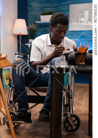Male drawing professional wheelchair user writing notes with paper and pencil on desk in bedroom. African american artist living with medical condition making hand drawn sketch with colored pencil. Male drawing professional wheelchair user writing notes with paper and pencil on desk in bedroom. African american artist living with medical condition making hand drawn sketch with colored pencil. 96706830