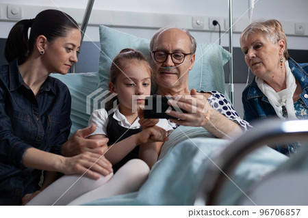 Young woman, daughter showing how cell phone works to sick grandfather at hospital observation room. Family watching smartphone movies at elderly people medical clinic rest room. 96706857