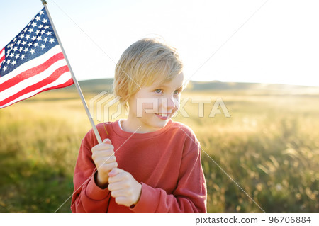 Cute little boy celebrating of July, 4 Independence Day of USA at sunny summer sunset. Child running and jumping with american flag symbol of United States over wheat field. Patriotism 96706884