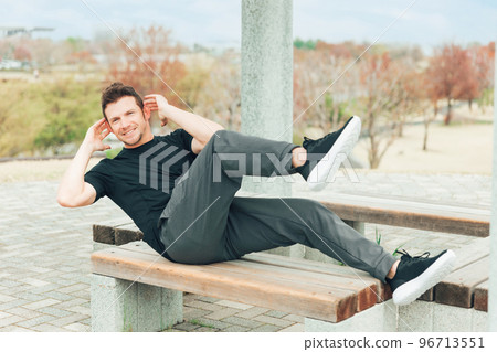 Caucasian man in sportswear doing twist crunches and abdominal muscles on a park bench Caucasian man in sportswear doing twist crunches and abdominal muscles on a park bench 96713551