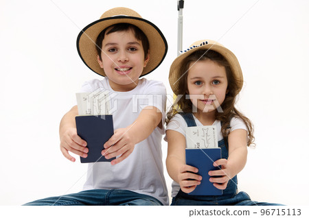 Happy journey! Family of travelers brother and sister wearing casual denim, white t-shirt and straw hat, with suitcase, showing on camera air tickets and passports, isolated on white background Happy journey! Family of travelers brother and sister wearing casual denim, white t-shirt and straw hat, with suitcase, showing on camera air tickets and passports, isolated on white background 96715113