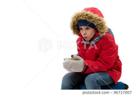 Charming preteen traveler boy in red down jacket and wool mittens, going for vacations, warms his hands with cup of hot drink, sits on suitcase, waits to board a flight. Travelling at winter holidays Charming preteen traveler boy in red down jacket and wool mittens, going for vacations, warms his hands with cup of hot drink, sits on suitcase, waits to board a flight. Travelling at winter holidays 96717007