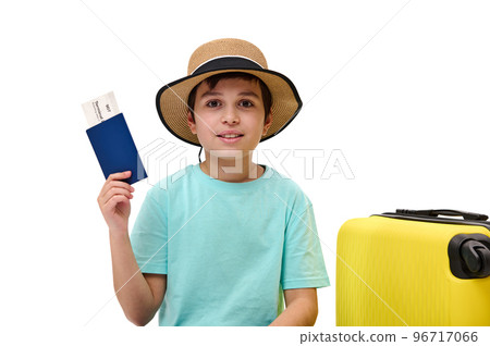Adorable preteen traveler boy wearing blue t-shirt and straw hat, going for summer vacations, shows boarding pass to camera, cutely smiles sitting near yellow suitcase, isolated over white background Adorable preteen traveler boy wearing blue t-shirt and straw hat, going for summer vacations, shows boarding pass to camera, cutely smiles sitting near yellow suitcase, isolated over white background 96717066