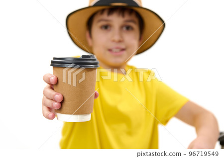 Selective focus on a disposable cardboard cup with takeaway hot drink in the hands of a blurred teenage boy in yellow t-shirt and straw hat, handing paper mug on camera, isolated over white background Selective focus on a disposable cardboard cup with takeaway hot drink in the hands of a blurred teenage boy in yellow t-shirt and straw hat, handing paper mug on camera, isolated over white background 96719549