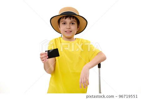 Isolated portrait on white background of a Latin American happy teenage traveler boy in yellow t-shirt and straw hat, with suitcase, showing a blank black plastic card with free ad space on camera 96719551