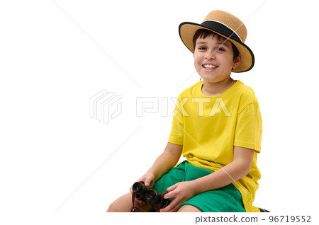 Cheerful multi-ethnic preteen boy, young traveler tourist in bright yellow t-shirt, green shorts and straw hat, with binoculars, sitting on suitcase and looking at camera, isolated on white background 96719552