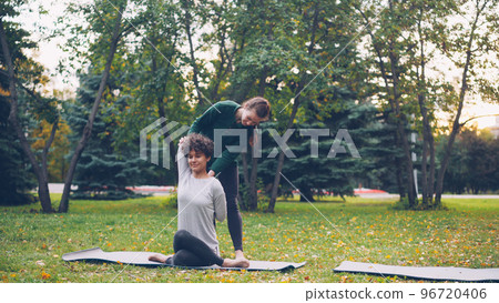Diligent yoga student is learning Cow Face position sitting on mat and stretching arms while experienced teacher is helping her, girls are talking and laughing at outdoor ptactice. 96720406
