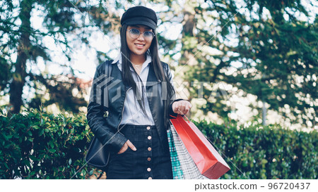 Portrait of attractive smiling Asian girl shopaholic standing outdoors in the street with shopping bags and looking at camera. Green trees and bushes are in background. Portrait of attractive smiling Asian girl shopaholic standing outdoors in the street with shopping bags and looking at camera. Green trees and bushes are in background. 96720437