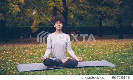 Good-looking girl in sports clothing is twisting her body sitting in lotus position then relaxing with closed eyes after practice in town park on autumn day. 96720448