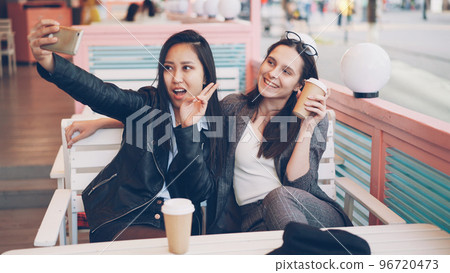 Carefree girls are taking selfie with take out drinks sitting in cafe together and using smartphone. Young women are posing, clinking glasses and showing hand gestures v-sign. Carefree girls are taking selfie with take out drinks sitting in cafe together and using smartphone. Young women are posing, clinking glasses and showing hand gestures v-sign. 96720473