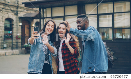 Excited young people foreigners are making online video call using smartphone holding gadget and talking showing building behind them expressing positive emotions showing thumbs-up. 96720477