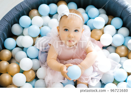 A cute one-year-old baby in a pink dress is bathing in a toy pool with colorful balloons. She is holding two balloons A cute one-year-old baby in a pink dress is bathing in a toy pool with colorful balloons. She is holding two balloons 96721680