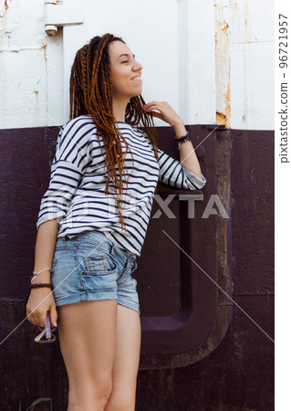 a girl with a dreadlocked hairstyle poses in the summer outdoors, dressed in a T-shirt and denim shorts 96721957