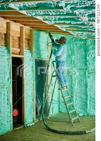 Male builder insulating wooden frame house. Man worker spraying polyurethane foam inside of future cottage, standing on ladder, using plural component gun. Construction and insulation concept. Male builder insulating wooden frame house. Man worker spraying polyurethane foam inside of future cottage, standing on ladder, using plural component gun. Construction and insulation concept. 96722139