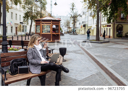 Business adult caucasian woman sitting with laptop and talking on speakerphone in city square Business adult caucasian woman sitting with laptop and talking on speakerphone in city square 96723705