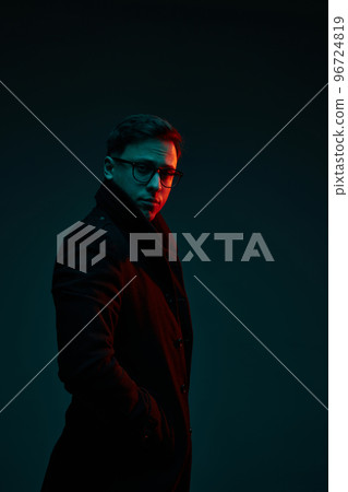 Studio shot of serious young 30s years old man in classic style clothes posing isolated over dark background in neon light. Fashion, style, emotions concept 96724819