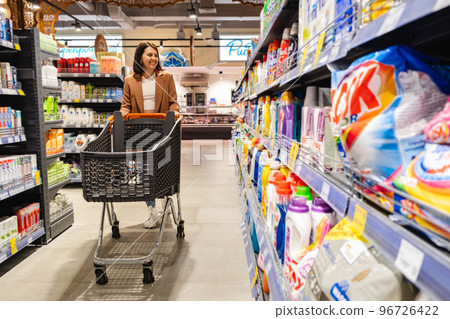 a woman with a cart walks between rows of shelves in a grocery store 96726422