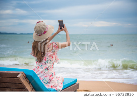 Summer beach vacation holidays trip concept, Happy young Asian woman with hat relaxing and taking a photo and selfie with smartphone on beach chair. 96728930