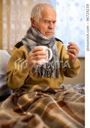 gray haired elderly man looking at body temperature on thermometer at home 96729239