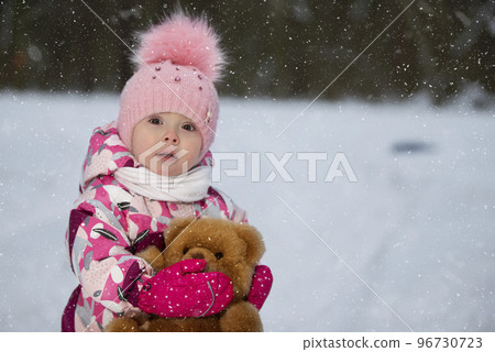 Little girl in winter with a teddy bear on the background of falling snow. Child on a winter walk. 96730723
