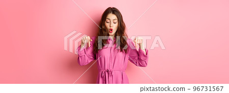 Beautiful female model in spring outfit, pointing and looking down, showing copyspace aside, standing against pink background 96731567