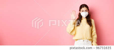 Covid-19, lockdown and pandemic concept. Young woman wearing face mask during quarantine, saying hello, waving raised hand to greet person from distance, pink background 96731635