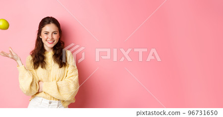 Young smiling woman looking with confidence, throwing apple in air, eating healthy fruits to keep that smile perfect, standing against pink background 96731650