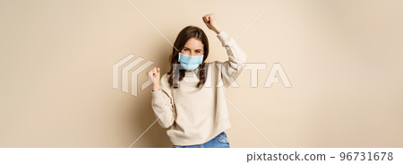 Covid-19, pandemic and quarantine concept. Enthusiastic girl dancing in face medical mask, celebrating, standing over beige background Covid-19, pandemic and quarantine concept. Enthusiastic girl dancing in face medical mask, celebrating, standing over beige background 96731678