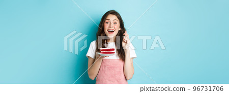 Holidays and celebration. Excited woman celebrating birthday, cross fingers and making wish while blowing candle on cake, standing against blue background 96731706