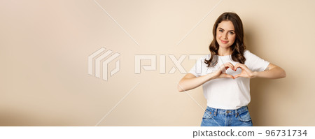 Lovely woman showing heart, love sign and smiling, standing over beige background in white t-shirt, beige background 96731734