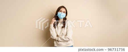 Covid-19, pandemic and quarantine concept. Beautiful modern woman in medical face mask, cross arms on chest, looking confident, beige background 96731736