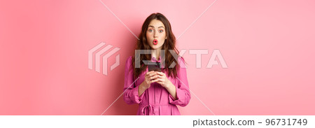 Image of surprised young woman gasping and saying wow, staring impressed at camera after reading news online on mobile phone, standing with smartphone against pink background Image of surprised young woman gasping and saying wow, staring impressed at camera after reading news online on mobile phone, standing with smartphone against pink background 96731749