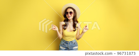 Stylish girl in straw hat and tank top, ready for summer, going on vacation and smiling pleased, standing over yellow background 96731873