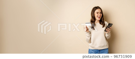 Smiling girl using mobile app, smartphone shopping and credit card, standing over beige background, order smth Smiling girl using mobile app, smartphone shopping and credit card, standing over beige background, order smth 96731909
