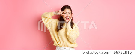 Attractive female model, gasping amazed, showing v-sign over eye, smiling happy, standing against pink background Attractive female model, gasping amazed, showing v-sign over eye, smiling happy, standing against pink background 96731910