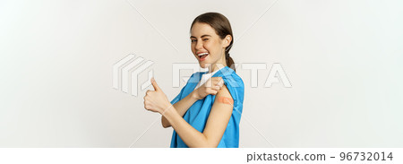 Smiling nurse, doctor in scrubs, medical worker showing her vaccinated shoulder with patch, thumbs up, recommending vaccinate from coronavirus, white background 96732014