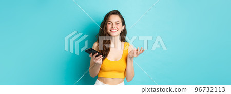 Technology and lifestyle concept. Beautiful caucasian woman chatting on phone, holding smartphone and smiling at camera, blue background 96732113