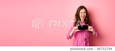 Technology concept. Cheerful lady in dress wink at you, smiling and showing mobile phone screen horizontally, standing over pink background 96732794