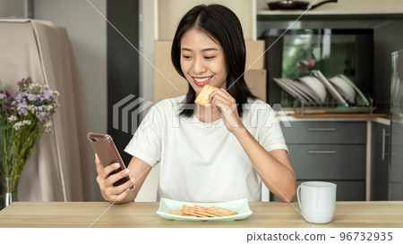 Asian woman eats crackers for breakfast and uses her mobile phone to check the morning news before going to work, Working people's urgent food, Urgent activity after waking up 96732935