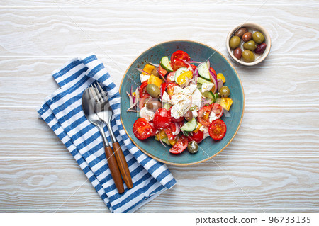 Greek salad with feta cheese, vegetables, olives in blue bowl on white wooden background Greek salad with feta cheese, vegetables, olives in blue bowl on white wooden background 96733135