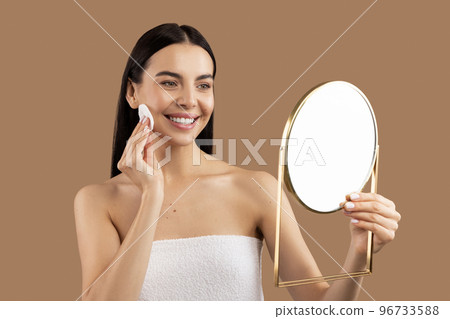 Brunette woman cleansing face with cotton pad, looking at mirror 96733588