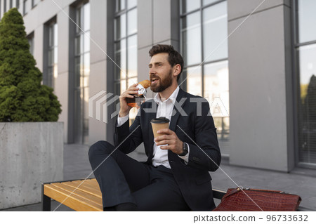 Happy hangry young european male manager with beard in suit with cup of coffee takeaway eats croissant 96733632