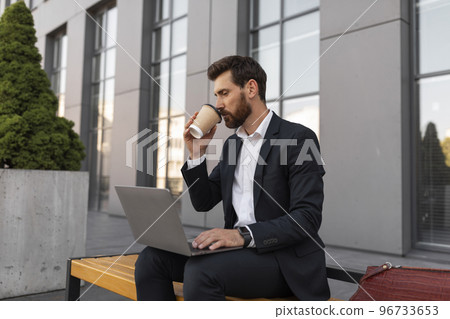 Happy busy young european man manager with beard in suit typing on computer, drink cup of coffee takeaway Happy busy young european man manager with beard in suit typing on computer, drink cup of coffee takeaway 96733653