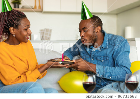 Loving african american couple in party hats celebrating birthday, man blowing candle on festive pie, sitting on sofa 96733668