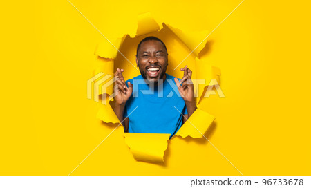 Wishing good luck. Emotional black man gesturing and crossing fingers, posing through hole in torn yellow paper Wishing good luck. Emotional black man gesturing and crossing fingers, posing through hole in torn yellow paper 96733678