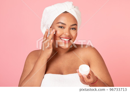 Beautiful african american body positive lady holding jar of cream, and applying moisturizer on cheek, pink background 96733713